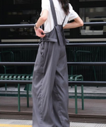 白いシャツにグレーのオーバーオールを着用し、後ろ姿でストラップを触る女性の全身写真。