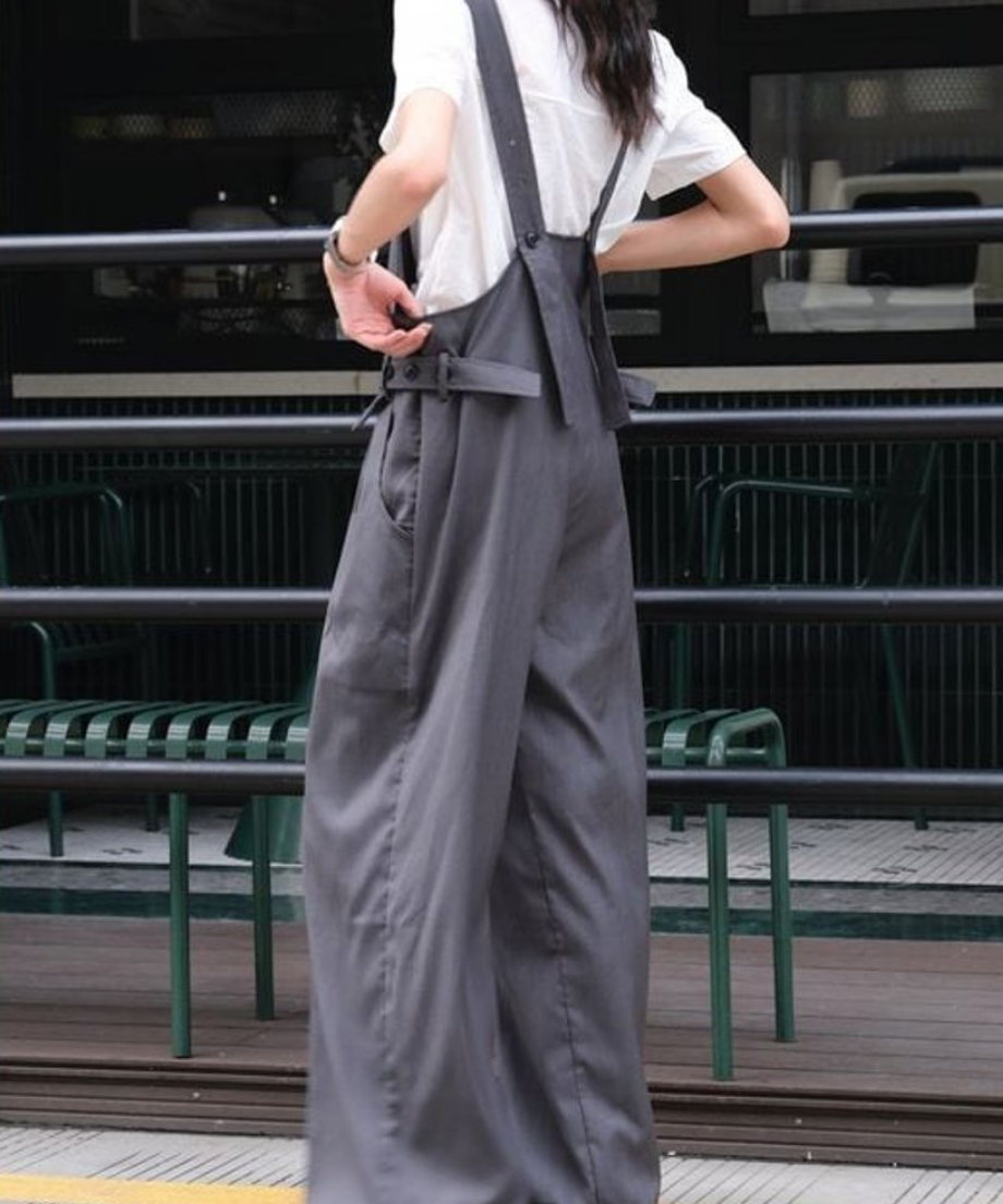 白いシャツにグレーのオーバーオールを着用し、後ろ姿でストラップを触る女性の全身写真。