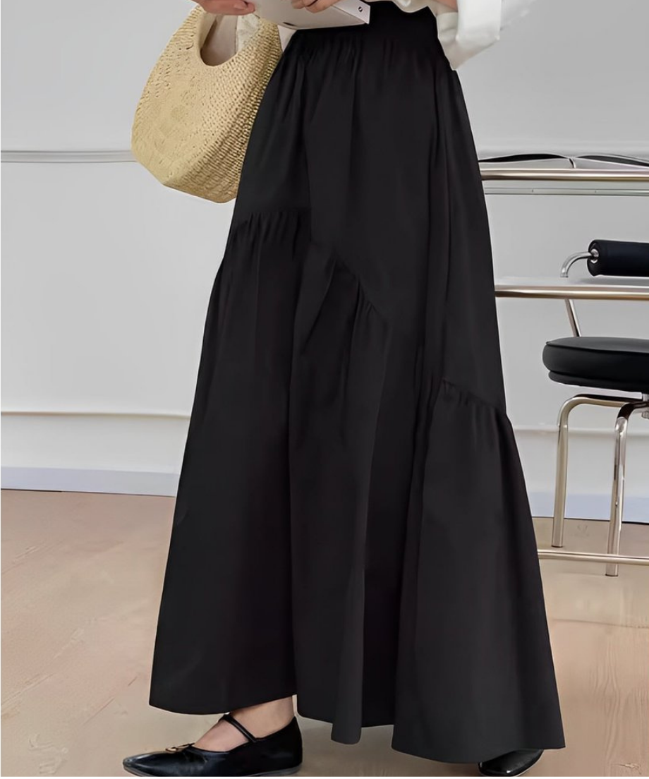 黒いティアード切り替えマキシスカートを履いた女性のアップ。白いトップスをインし、麦わら素材のバッグを持っている。椅子の前で撮影。