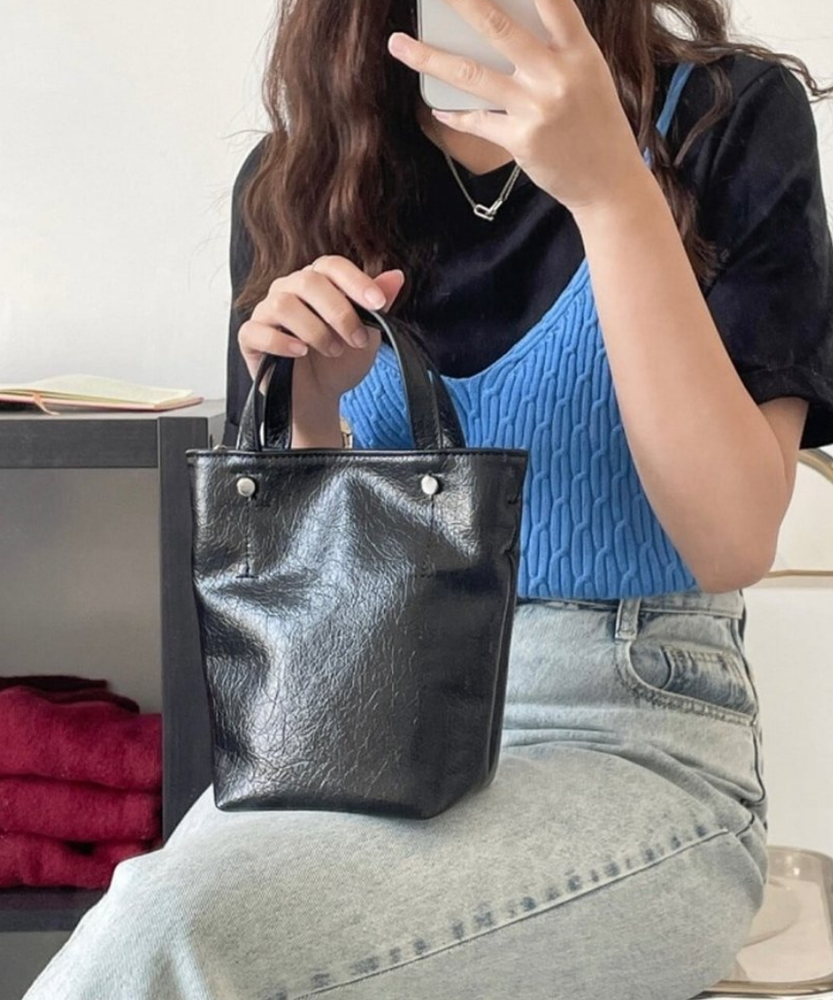 椅子に座り、膝の上に置いたブラックのミニトートバッグに手を添えるブルーのニットベストを着た女性