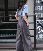 淡いブルーの半袖Tシャツにグレーのワイドレッグオーバーオールを着用し、横を向いて立つ女性の全身写真。