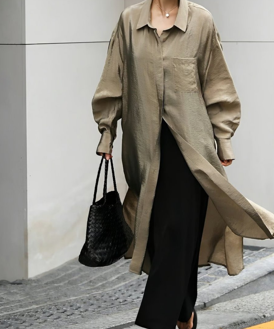 光沢のあるグレージュのロングシャツに黒のワイドパンツと編み込みバッグを合わせた、上品な外出スタイル。