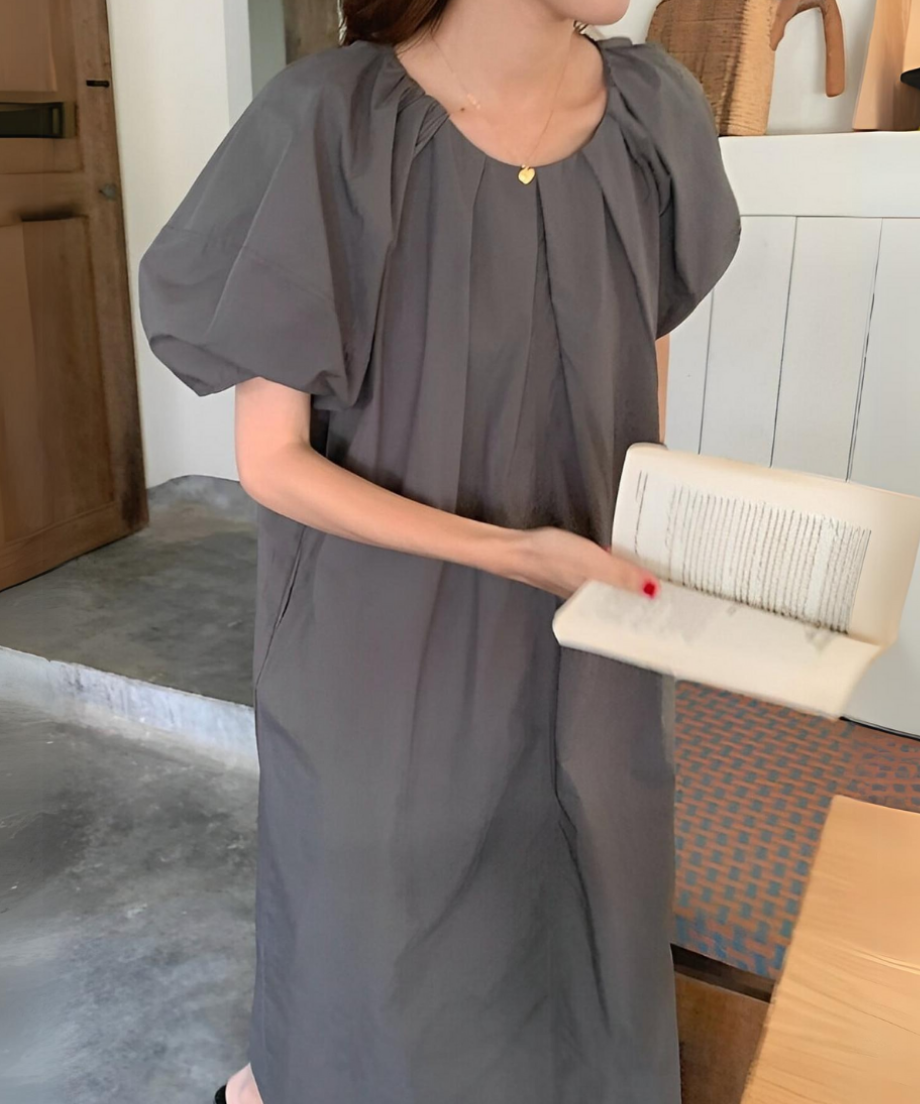 グレーのパフスリーブワンピースを着用し、本を手にする女性。首元のプリーツデザインが特徴。