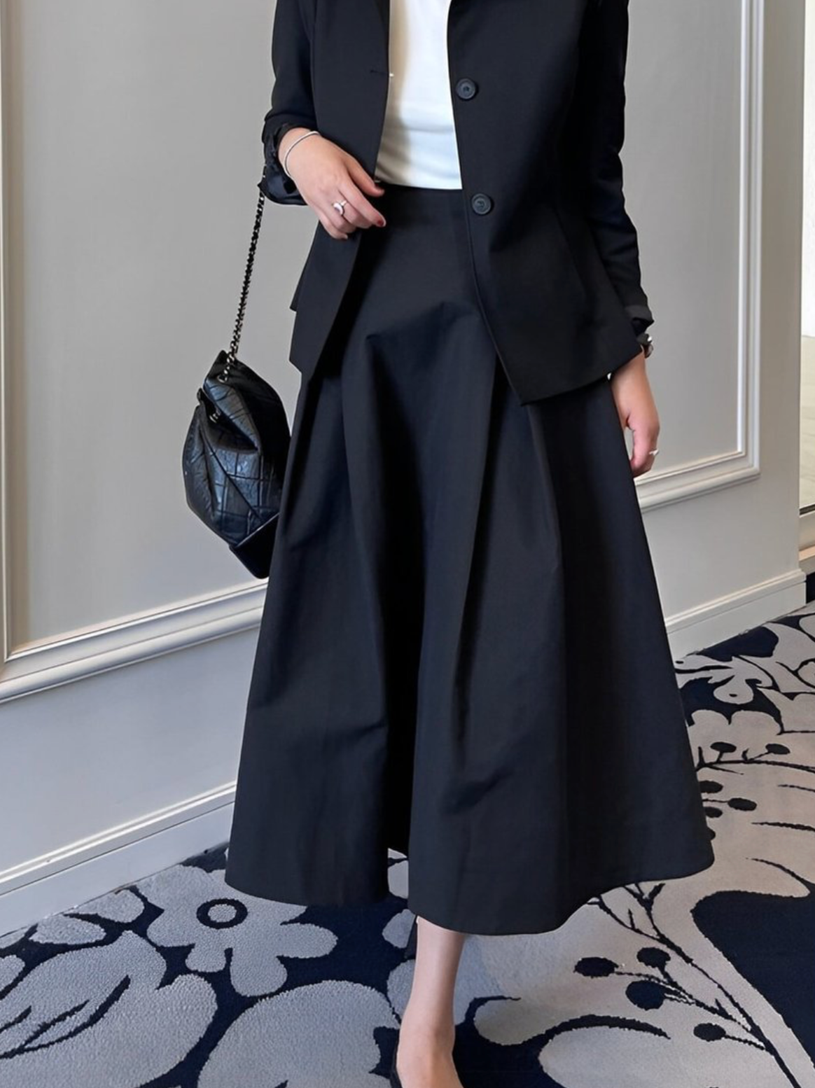 黒いジャケットと黒いミディ丈フレアスカートを着用した女性の半身。黒いキルティングバッグを腕にかけている。