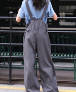 淡いブルーの半袖Tシャツにグレーのワイドレッグオーバーオールを着用し、後ろ向いて立つ女性の全身写真。