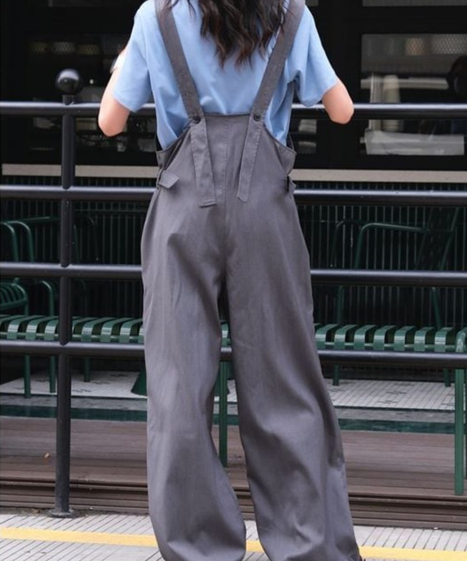 淡いブルーの半袖Tシャツにグレーのワイドレッグオーバーオールを着用し、後ろ向いて立つ女性の全身写真。