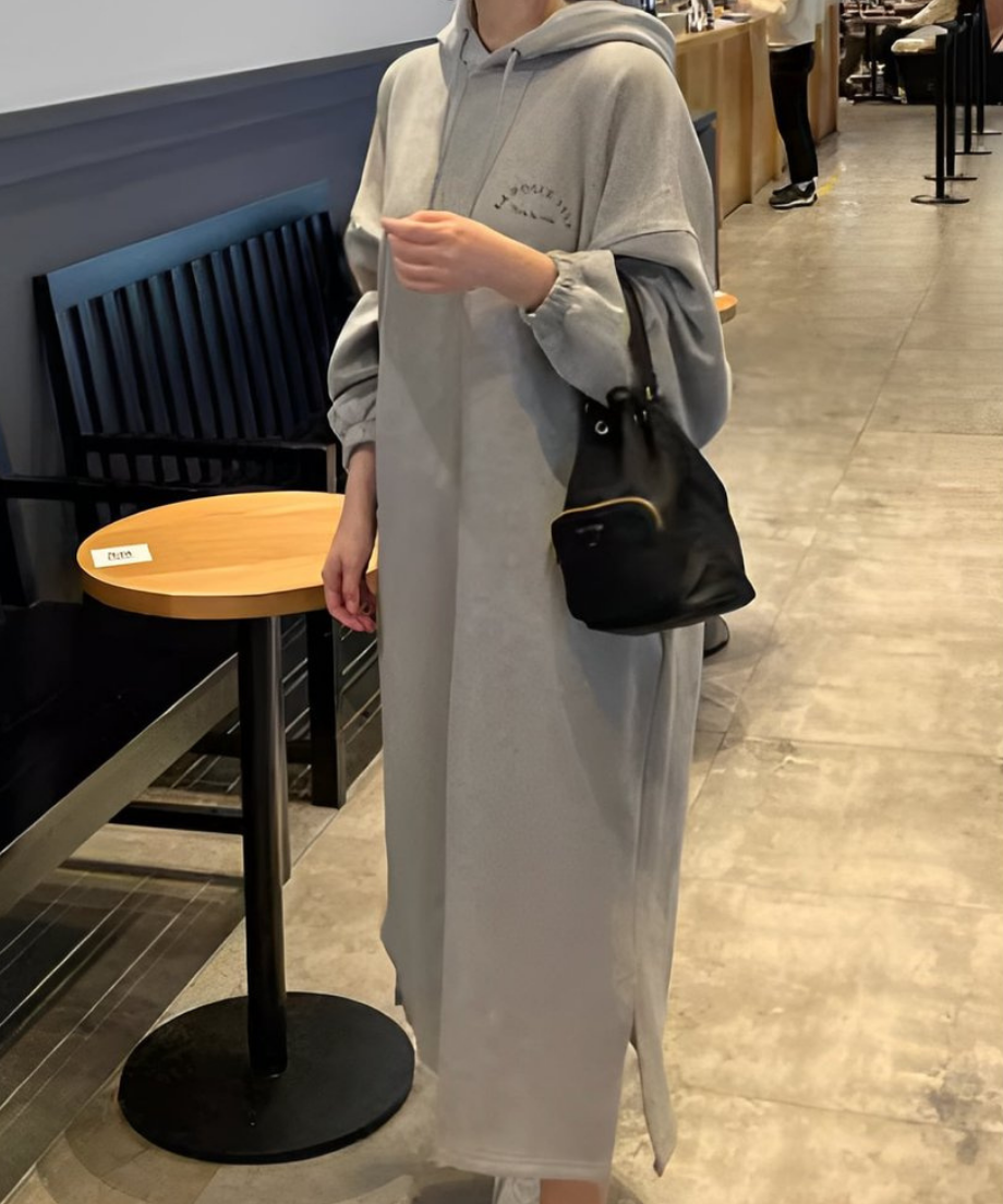 ロング丈フーディワンピースを着た女性が黒い巾着バッグを右手に持ち、オーバーサイズのゆったりとしたシルエットが特徴的。