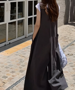 グレーのサロペットを着た女性が、柄物のバッグを持ち、屋外の建物前で立っている後ろ姿。
