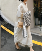 オフホワイトのロングシャツワンピースを着用し、ベージュのキルティングバッグを持ち、ボーダー柄のトップスを抱えて屋外を歩く女性。