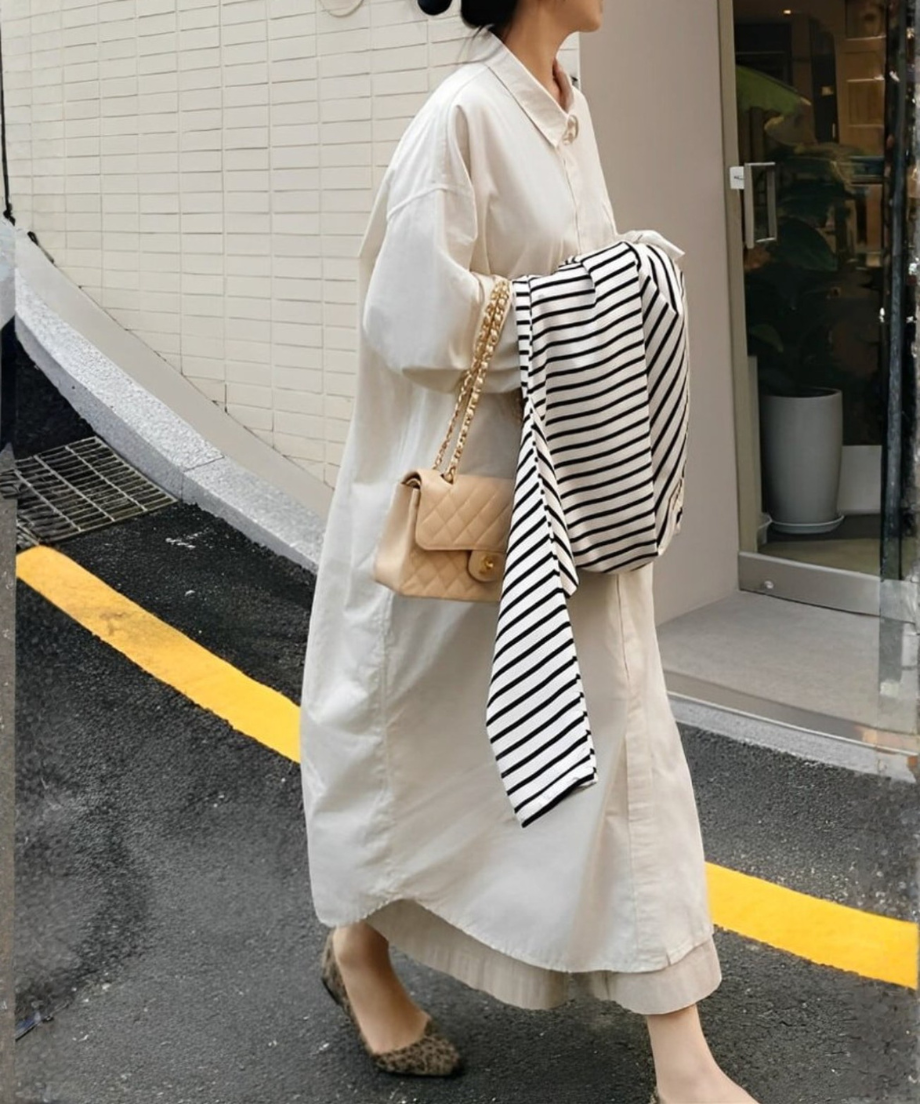 オフホワイトのロングシャツワンピースを着用し、ベージュのキルティングバッグを持ち、ボーダー柄のトップスを抱えて屋外を歩く女性。