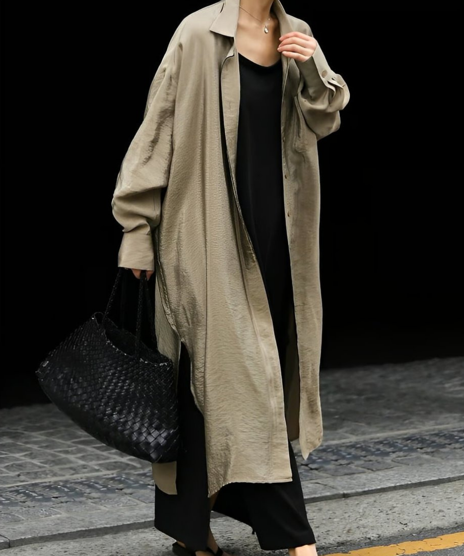 光沢のあるグレージュのロングシャツのバタンを全て開けて黒のワイドパンツと編み込みバッグを合わせたスタイル。