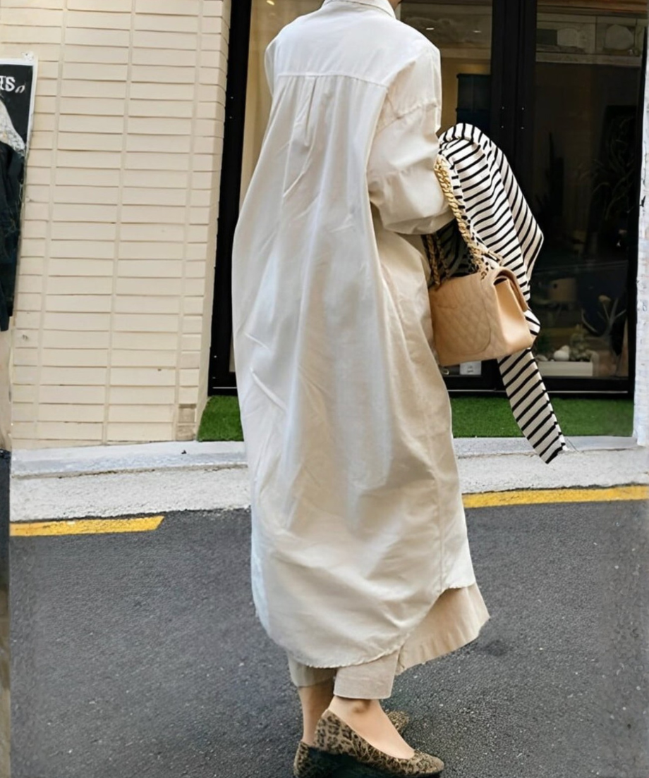 ロングシャツワンピースを着用し、ベージュのキルティングバッグを持ち、ボーダー柄のトップスを抱えて屋外を歩く女性。