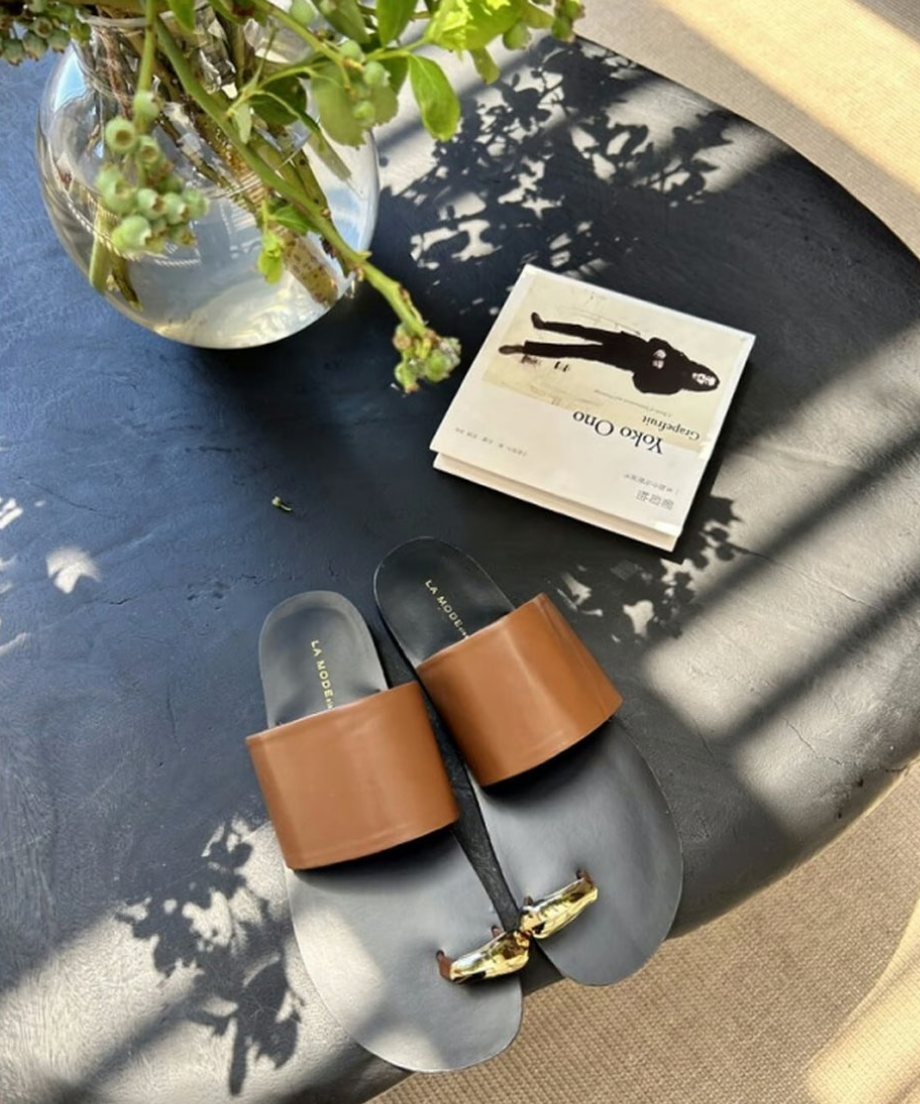 黒い円形のテーブルに置かれたブラウンのレザーサンダルと洋書、花瓶の風景