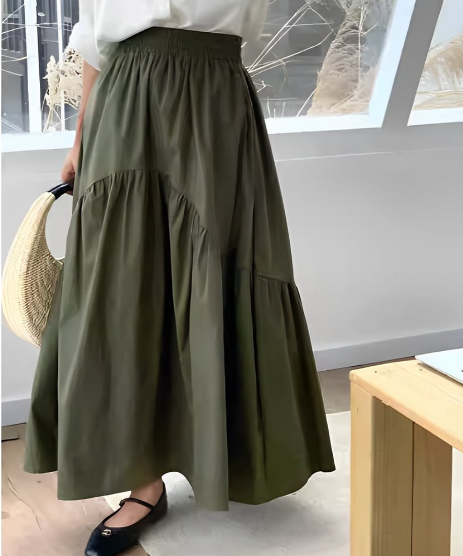 カーキ色のティアード切り替えマキシスカートを履いた女性のアップ。白いトップスをインし、麦わら素材のバッグを持っている。窓際で撮影。