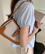 水色のパフスリーブブラウスを着た女性が、ブラウンのキャンバスバッグを肩にかけて本を持っている背後からのショット。