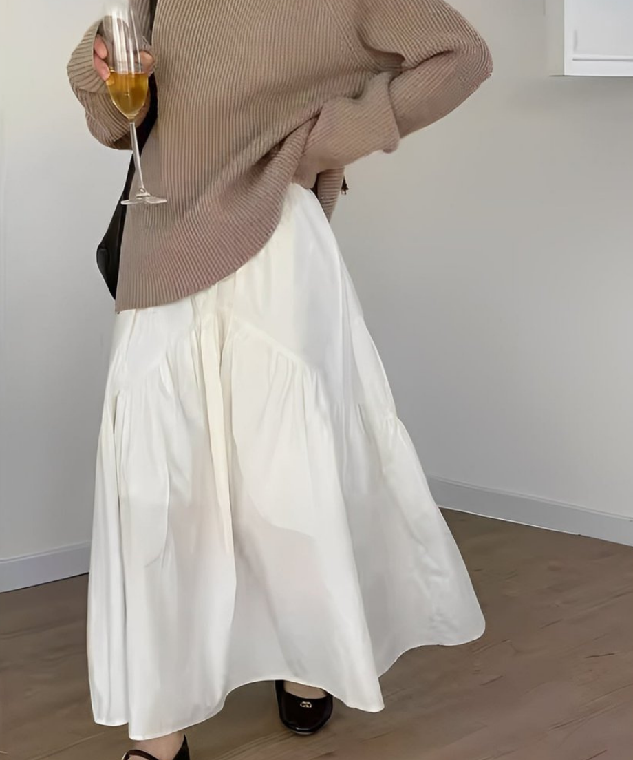 ホワイトのティアード切り替えマキシスカートを履いた女性が、ベージュの長袖ニットを着用し、黒いトートバッグを持っている全身ショット。
