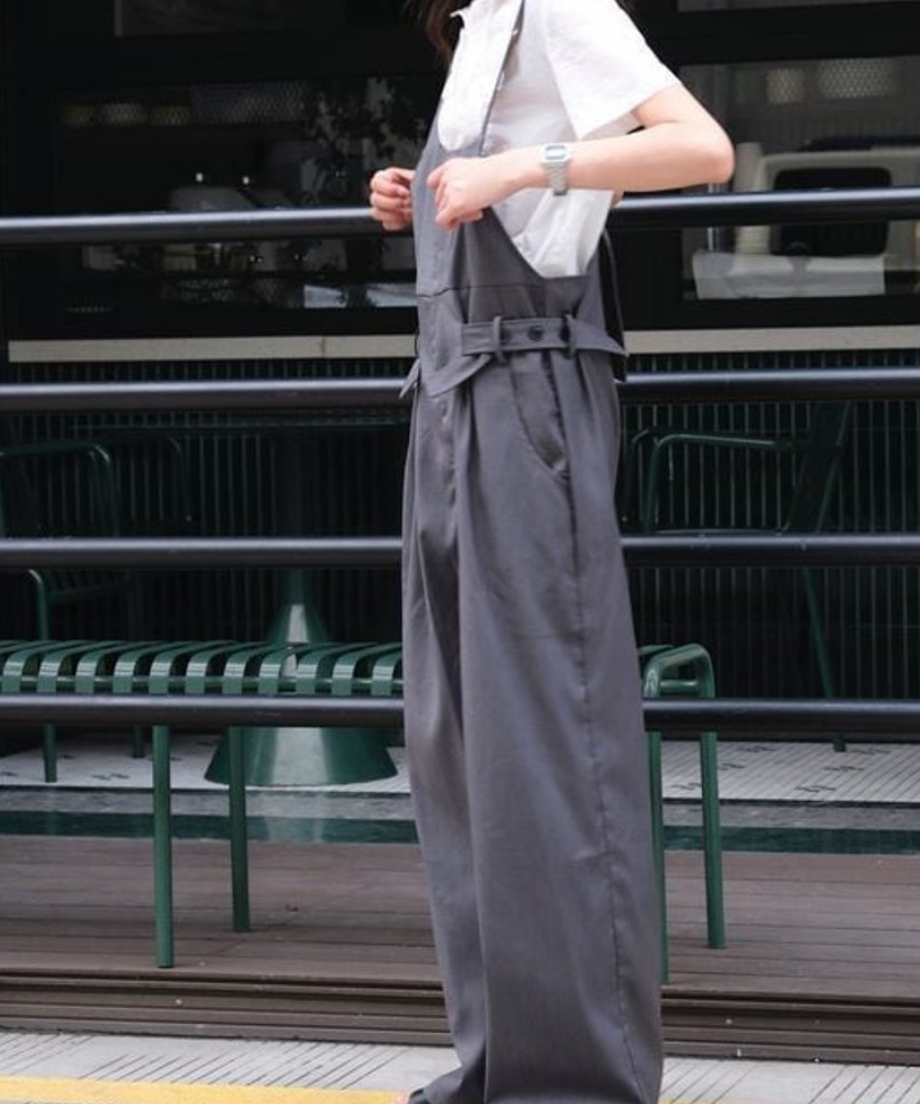 白いシャツにグレーのオーバーオールを着用し、横向き姿でストラップを触る女性の全身写真。
