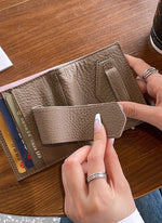 グレーのスリムな二つ折り財布を開き、内側のカードスロットとフラップの留め具部分を、ネイルをした手が操作している接写写真。