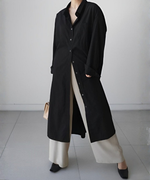 黒いロングシャツワンピースをボタンを３つ閉めて着用し、ベージュのパンツとキルティングのミニバッグを合わせた女性の正面