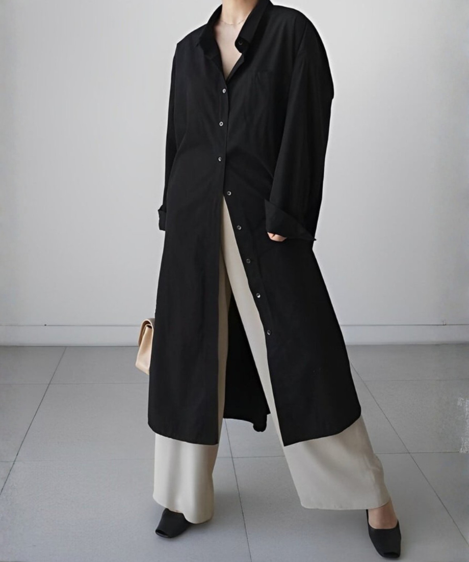 黒いロングシャツワンピースをボタンを３つ閉めて着用し、ベージュのパンツとキルティングのミニバッグを合わせた女性の正面