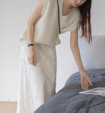 ホワイトのニットベストとホワイトの刺繍スカートを着た女性が、ベッドに置かれた本に手を伸ばしている。