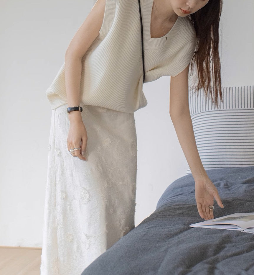 ホワイトのニットベストとホワイトの刺繍スカートを着た女性が、ベッドに置かれた本に手を伸ばしている。