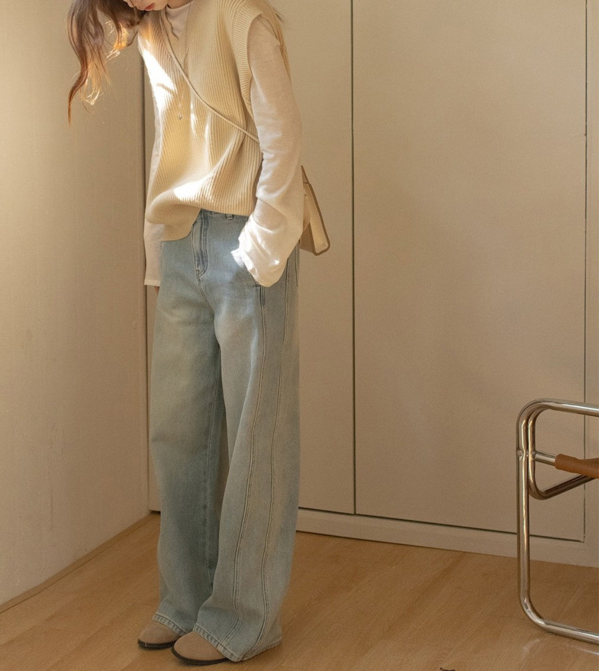 ベージュのニットベストとホワイトのロングスリーブ、ライトブルーのワイドレッグデニムを着た人物が部屋にいる。