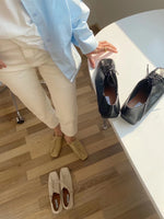 水色のシャツと白いパンツを着用した人物の足元と、白いテーブルの上に置かれた黒のスクエアトゥレースアップシューズのペア、床に置かれたアイボリーのシューズのペア。