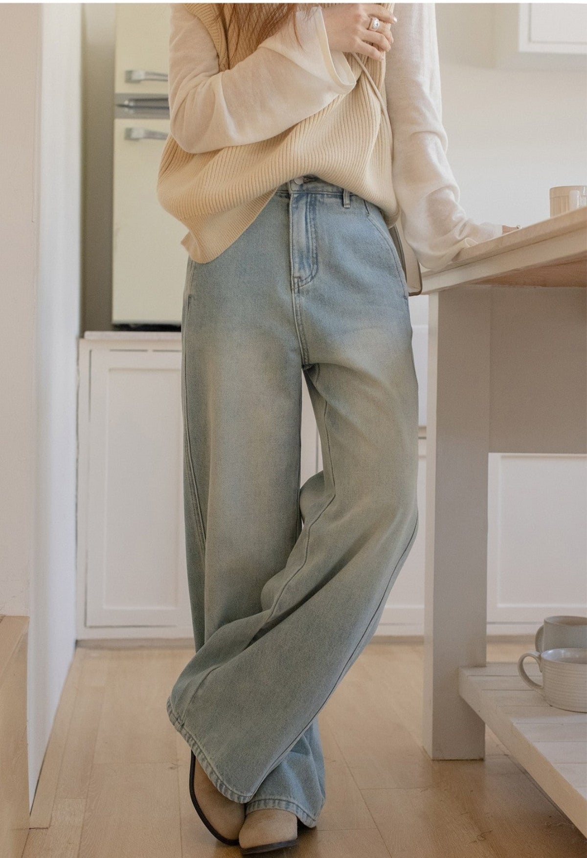 ライトブルーのワイドレッグデニムを履いたモデルが、キッチンでリラックスした姿勢で立っているシーン。