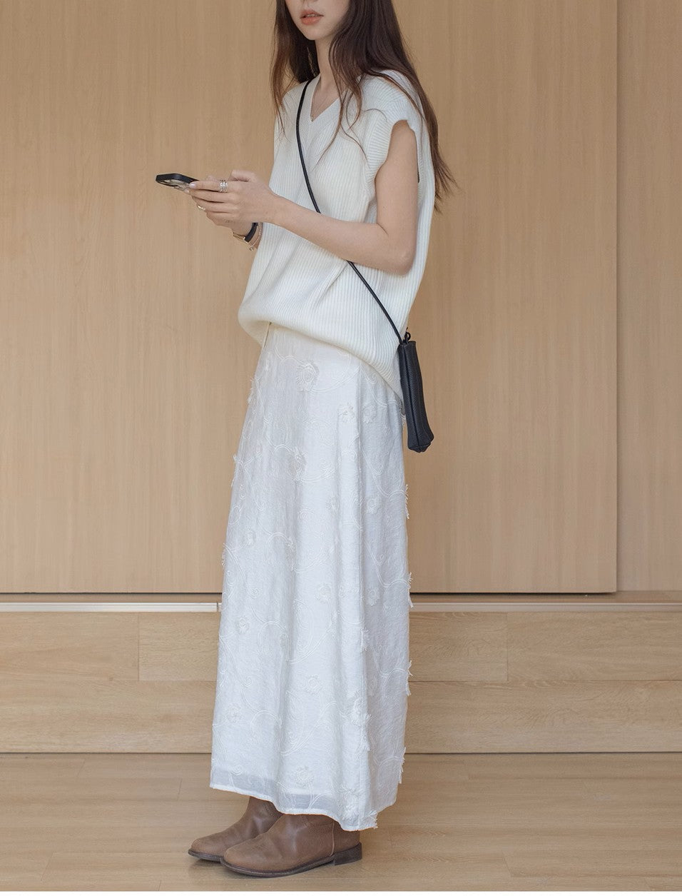 ニットベストとホワイトの刺繍スカートを着た女性が、スマートフォンを手に持ち、クロスボディバッグを身につけている。