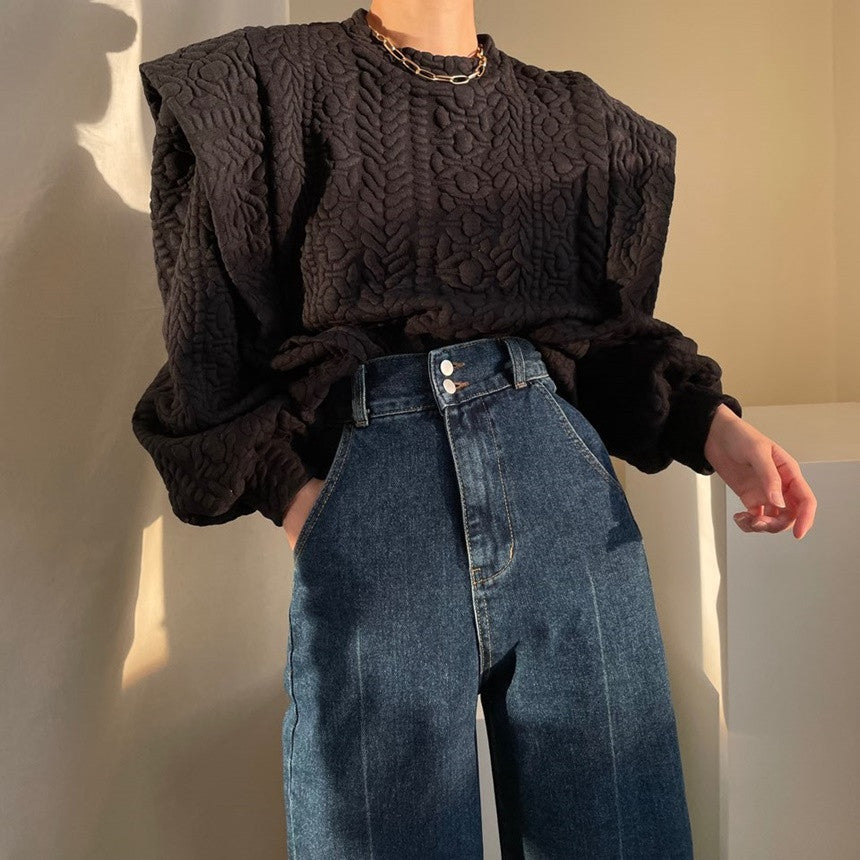 ブラックのエンボス加工素材プルオーバーを着用した女性の画像。特徴的なショルダーパッドと、濃いインディゴブルーのハイウエストワイドデニムを合わせている。