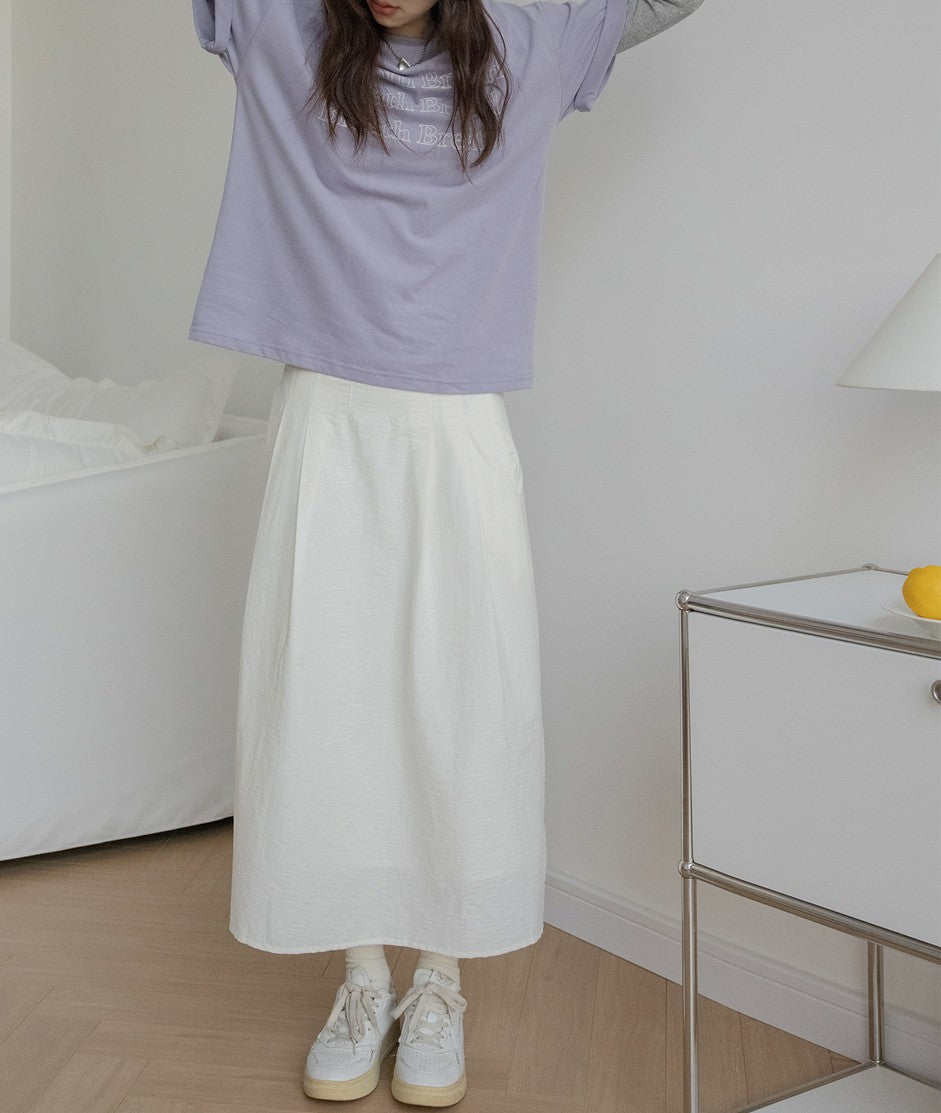 ラベンダー色のTシャツと白いプリーツスカートを着用した女性。リラックスしたスタイルでスニーカーを合わせている。