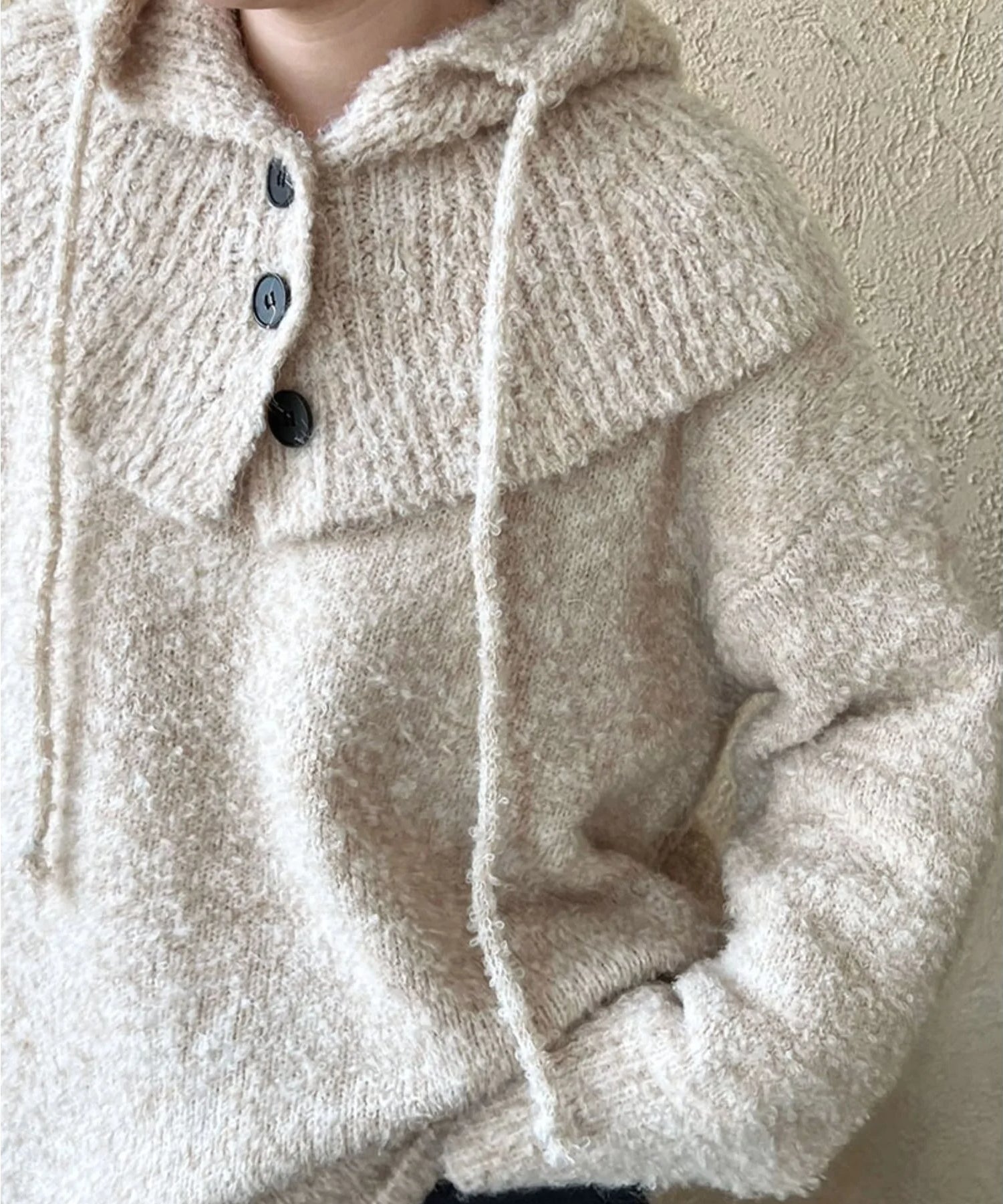 ベージュ色のざっくり編みクルーネックニットを着用した女性のバストアップ。黒の三つのボタンが見える。