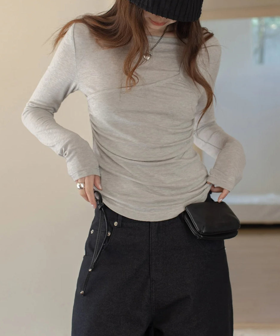 ライトグレーのレイヤードデザインのロングスリーブトップスを着用し、黒のボトムスを合わせた女性のクローズアップ。ドレープと袖口のサムホールが特徴的で、帽子をかぶったショット。