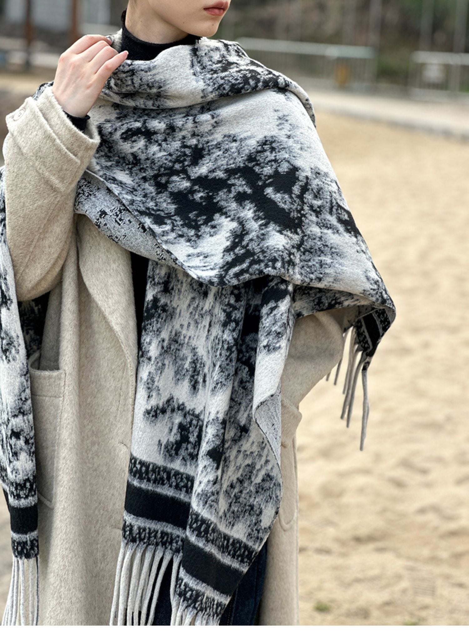 モノトーンの水墨画風の柄ショールをベージュのコートと合わせて屋外で着用する女性。