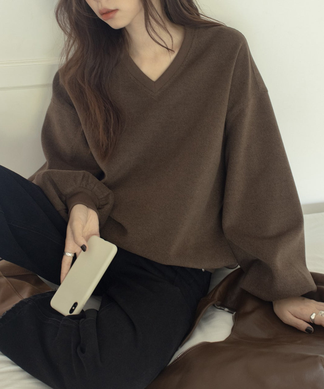 ブラウンのVネックバルーンスリーブトップスを着た女性のクローズアップ。手にスマホをもって座っている。