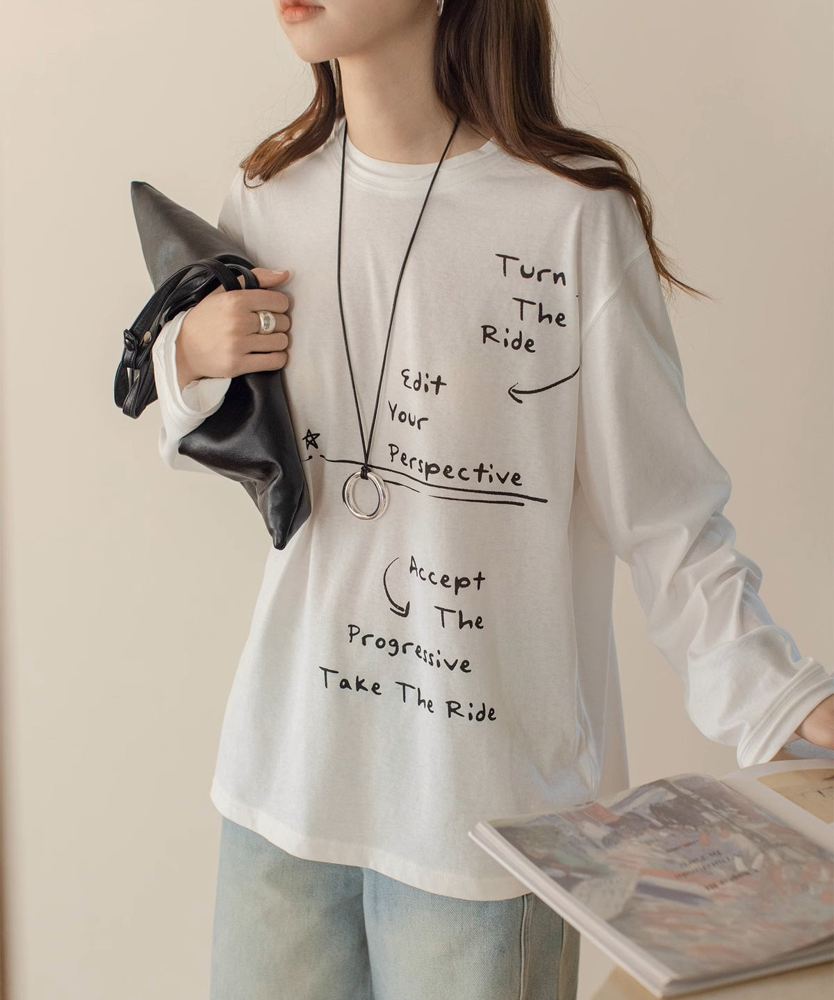 白のオーバーサイズロングTシャツを着用した女性の半身。Tシャツには手書き風の英文メッセージとイラストのプリントがあり、黒のレザー調バッグとロングネックレスを着用。