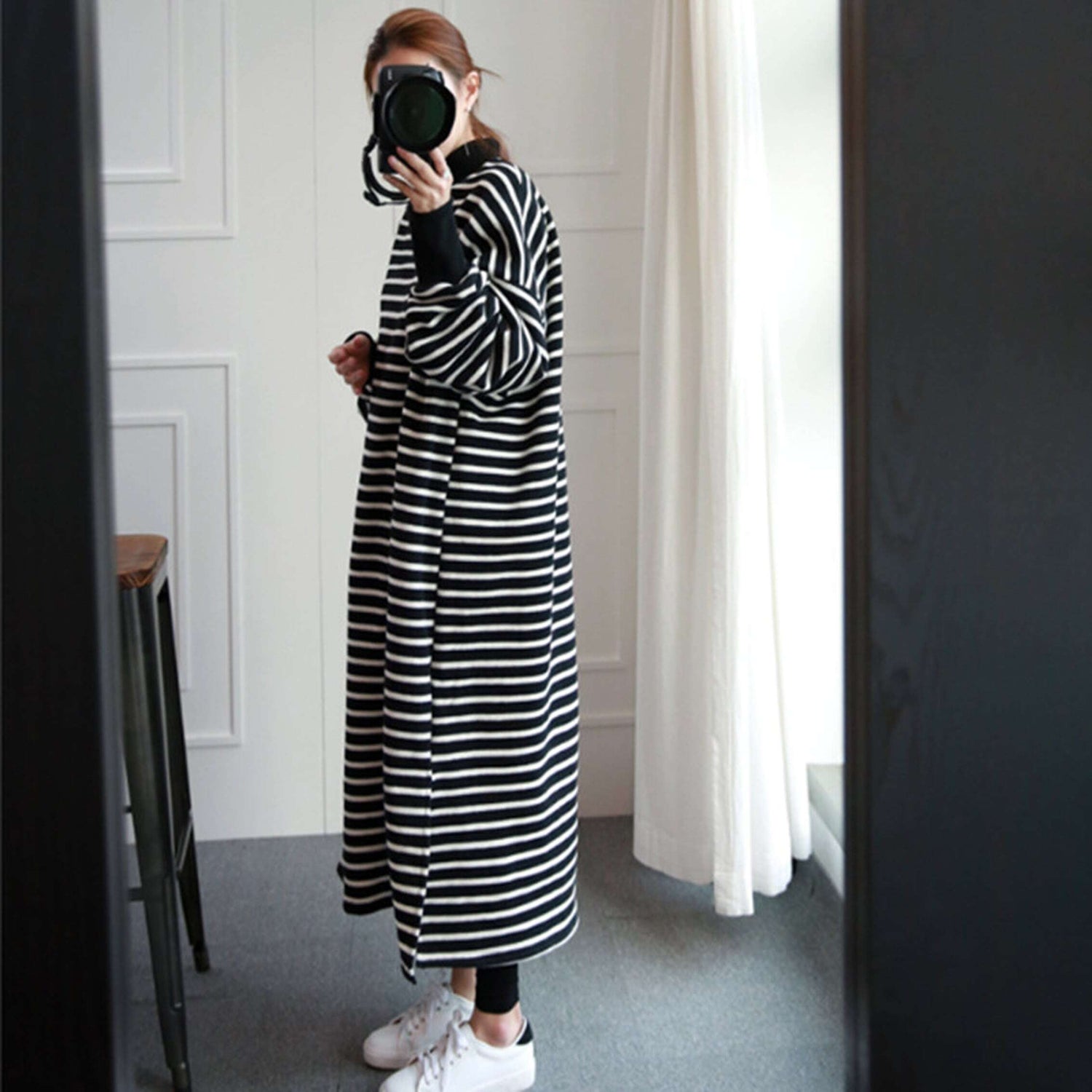 黒と白の細めボーダー柄ロングワンピースを着用し、黒いレギンスと白いスニーカーを履いた女性が、一眼レフカメラで自身を鏡越しに撮影している正面の全身写真。