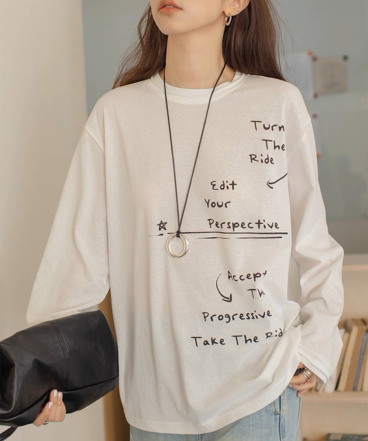 白のオーバーサイズロングTシャツを着用した女性の半身。Tシャツには手書き風の英文メッセージとイラストのプリントがあり、黒のレザー調クラッチバッグとロングネックレスを着用。