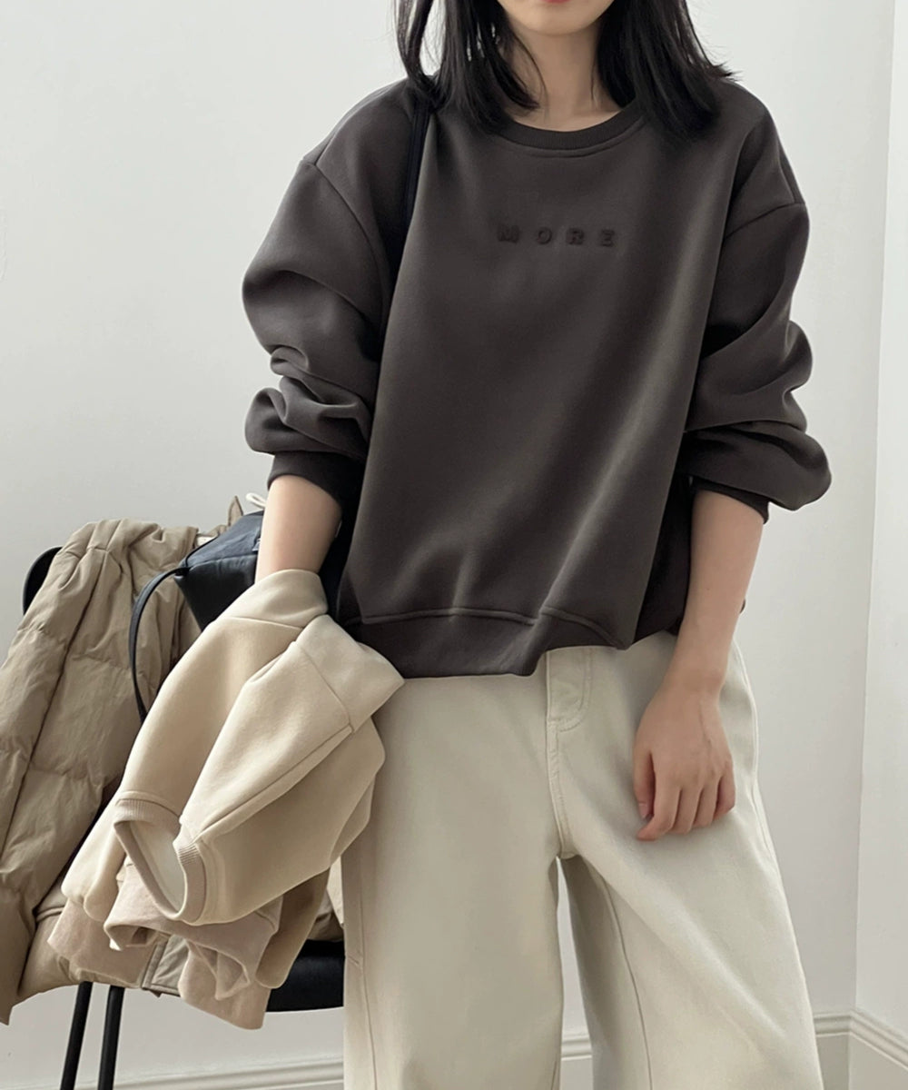 ダークブラウンのスウェットシャツを着た女性がベージュのスウェットを手にもって立っている様子。