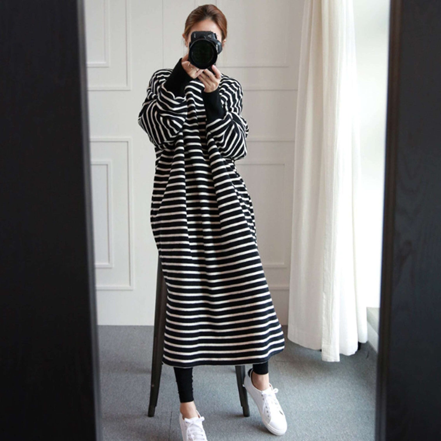 白黒ボーダー柄のロングワンピースを着て、白いスニーカーを履いた女性が、カメラで自身の姿を鏡越しに撮影している側面の立ち姿。