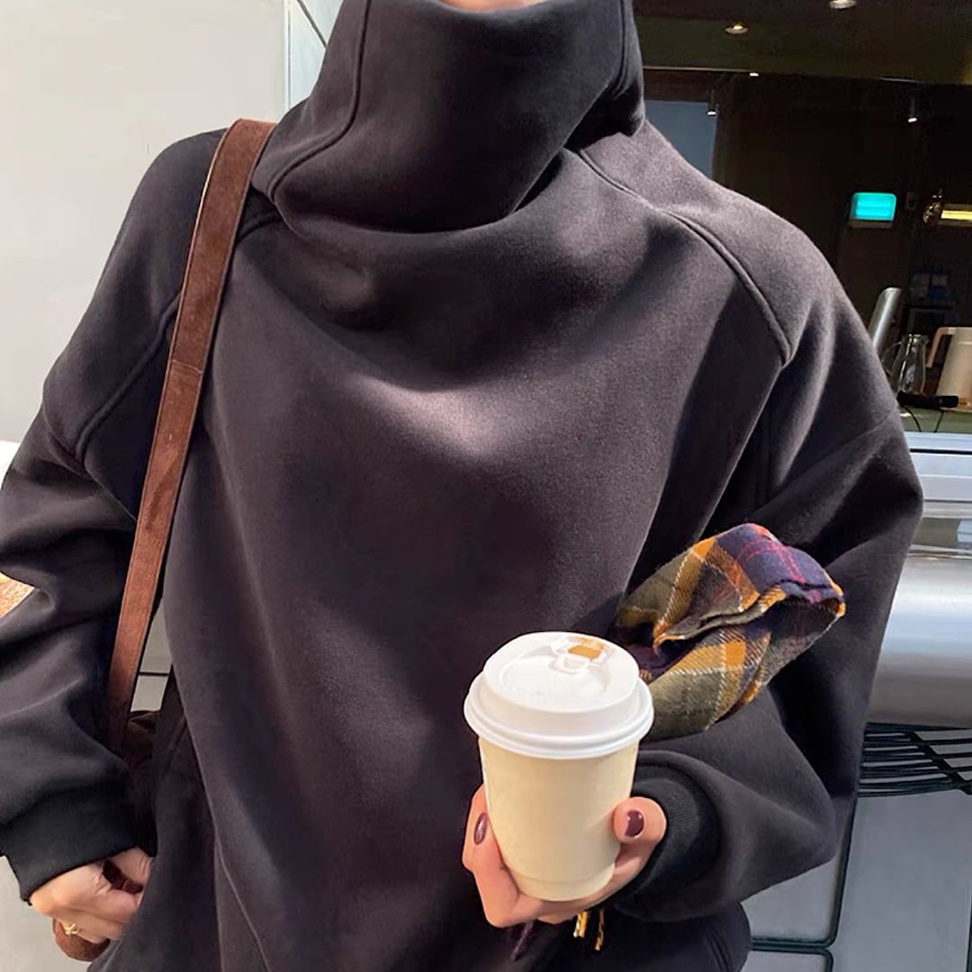 ブラックのタートルネックスウェットを着用し、コーヒーカップとチェック柄のポーチを持っている女性のバストアップ。
