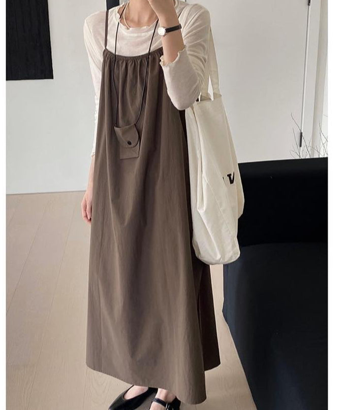 白い長袖トップスに茶色いロングキャミソールワンピースをレイヤードし、白いトートバッグを持つ女性の全身コーディネート。