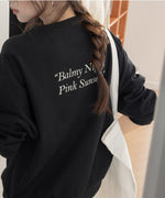 黒のオーバーサイズスウェットの背面クローズアップ。背中には「Balmy Night Pink Sunsets」の白色のプリントが施され、女性は白いトートバッグを肩にかけている。