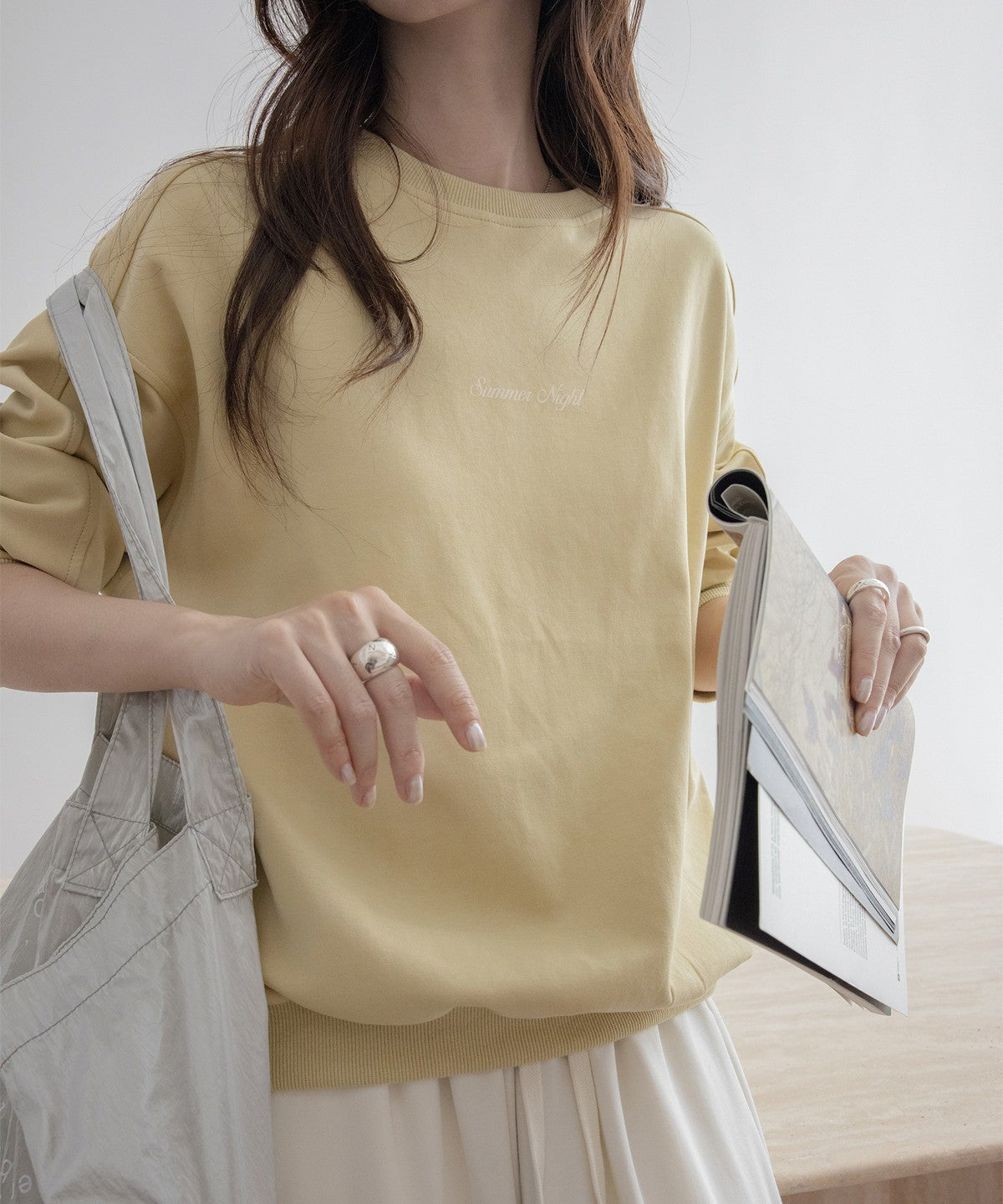 イエローのオーバーサイズスウェットと白のワイドパンツをコーディネートした女性。淡い色のショルダーバッグを持ち、手に本を持っている。