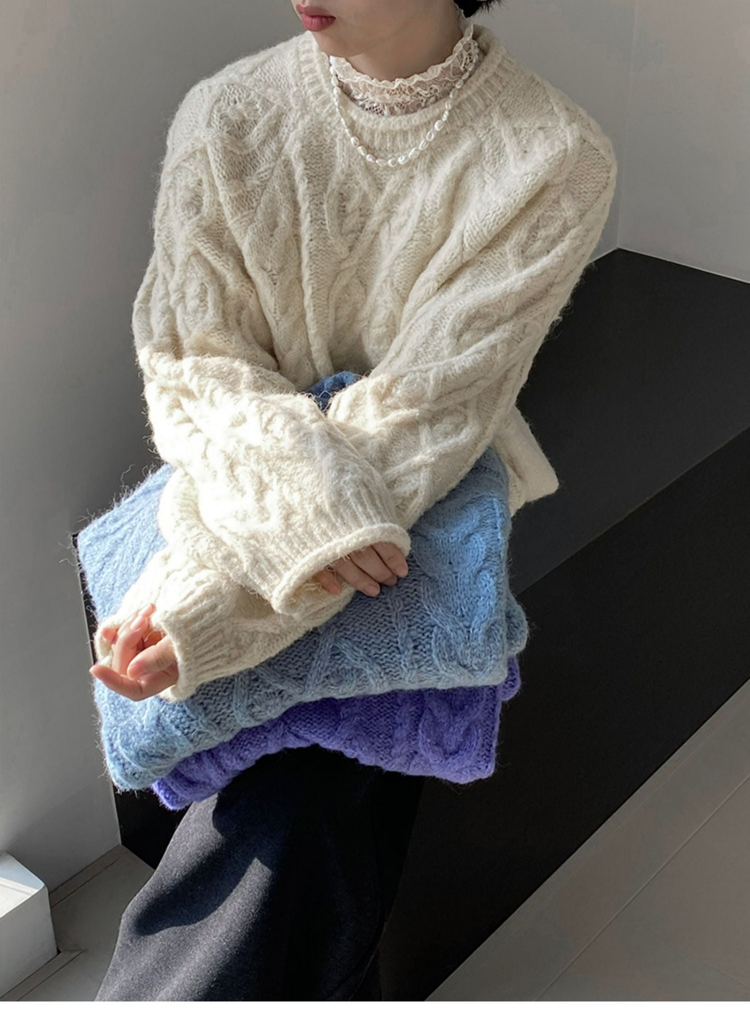 アプリコットのオーバーサイズケーブルニットを着用した女性が色違いのニットを膝にのせている。