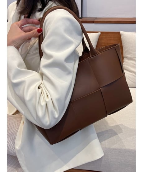 ホワイトのスーツを着た女性がダークブラウン色の編み込みデザインのトートバッグを肩にかけている写真