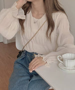 の細かいプリーツ加工シアーブラウスを着用し、濃い青のデニムを合わせた女性がテーブルについている。首元にはシンプルなペンダントネックレスを着用。