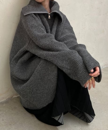 チャコールグレーのオーバーサイズハーフジップニットカーディガンを着用し、黒のプリーツスカートを合わせた女性の座りポーズ。