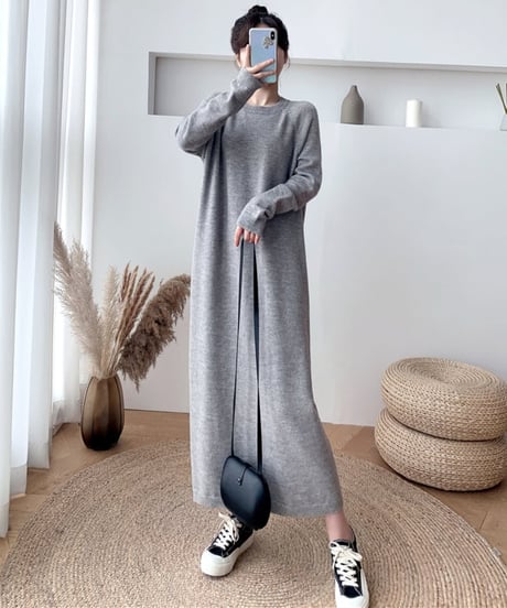 グレーの長袖ニットワンピースを着用し、黒いショルダーバッグを手に持っている女性の全身。